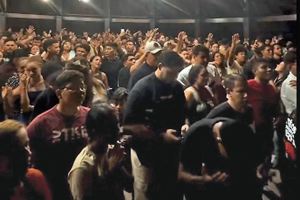 Centenas de pessoas reunidas em ambiente interno com teto de estrutura metálica e iluminação artificial. Público diversificado está de pé, muitos com as mãos levantadas, sugerindo participação ativa em evento.