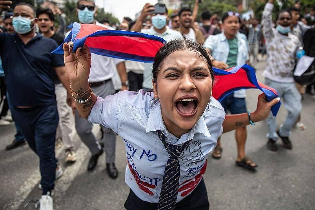 Jovem mulher no centro da imagem segura uma faixa azul e vermelha atrás da cabeça enquanto grita. Ao redor, grupo de pessoas caminha na rua, algumas usando máscaras, em manifestação pública durante o dia.