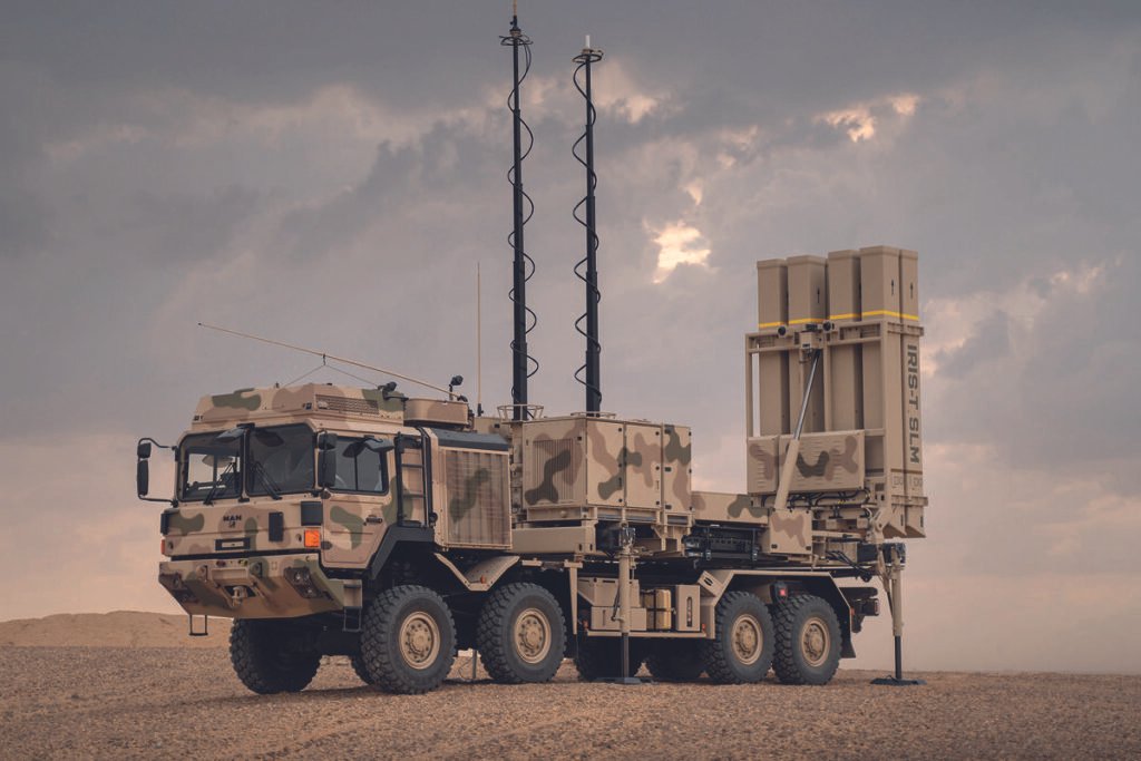 Veículo militar camuflado em tons de bege e verde com seis rodas estacionado em terreno árido sob céu nublado. O veículo possui duas antenas altas e um sistema de lançamento de mísseis na parte traseira