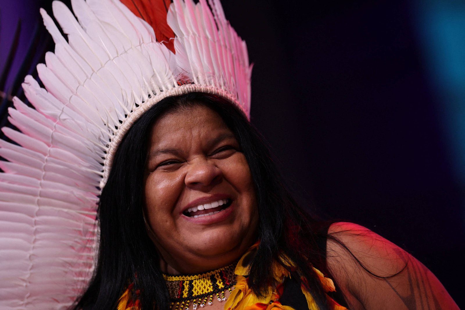 Mulher indígena sorrindo, usando cocar grande de penas brancas com detalhes vermelhos. Ela veste colar colorido e roupa com penas amarelas, com fundo escuro desfocado.