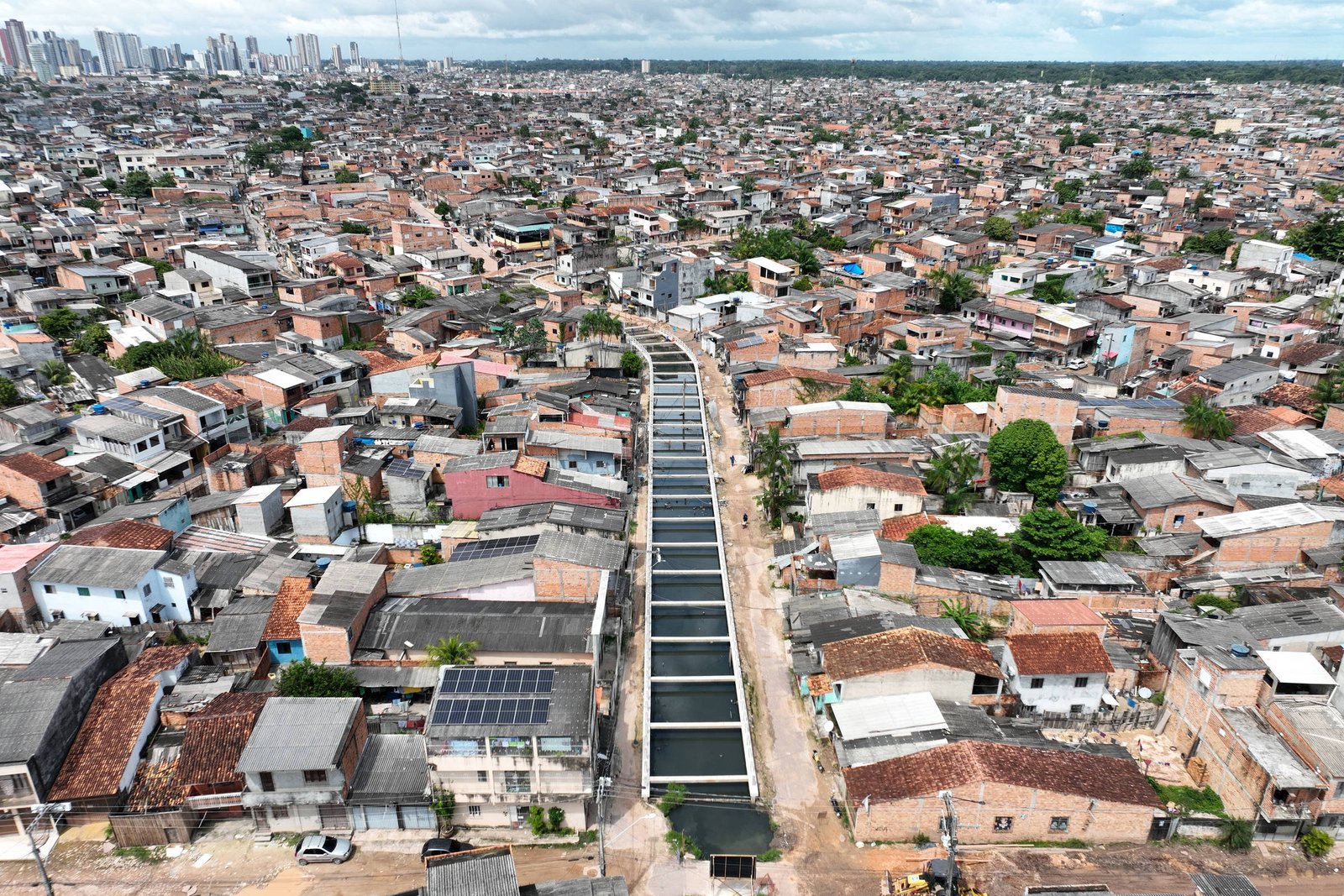 A imagem mostra uma vista aérea de uma área urbana densamente povoada, com muitas casas de diferentes tamanhos e cores. No centro, há uma via que parece ser um canal ou uma rua, ladeada por casas. Ao fundo, é possível ver uma cidade com prédios altos, indicando a proximidade de uma área mais urbanizada. O céu está parcialmente nublado.