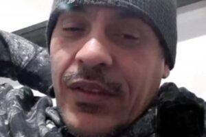 Homem de meia-idade com gorro cinza e jaqueta escura, com olhos parcialmente fechados, em ambiente interno com parede branca ao fundo.