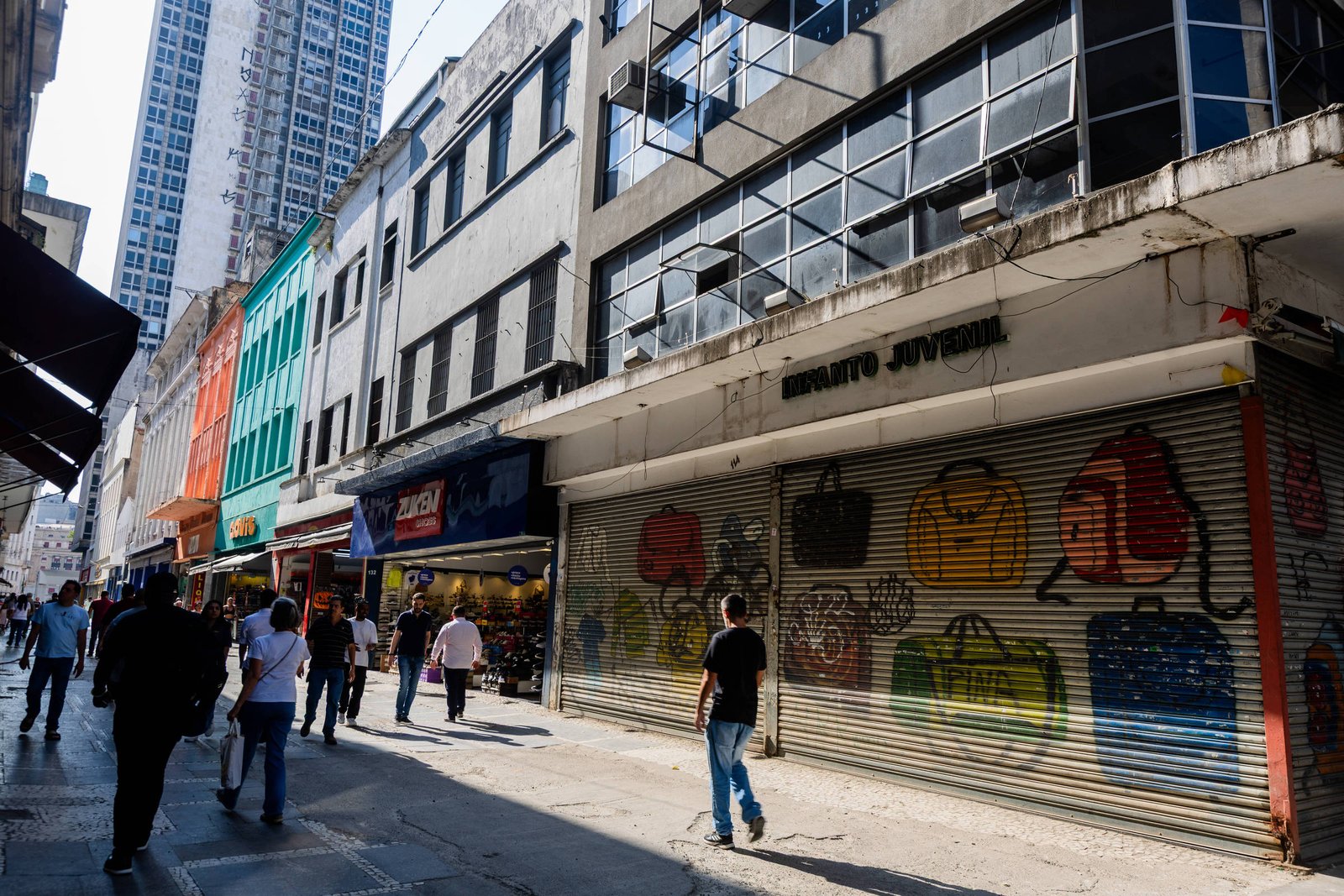 Rua movimentada com várias lojas de fachadas coloridas e pessoas caminhando. Uma loja com a porta metálica fechada exibe grafite de bolsas coloridas. Prédios altos aparecem ao fundo sob luz do dia.