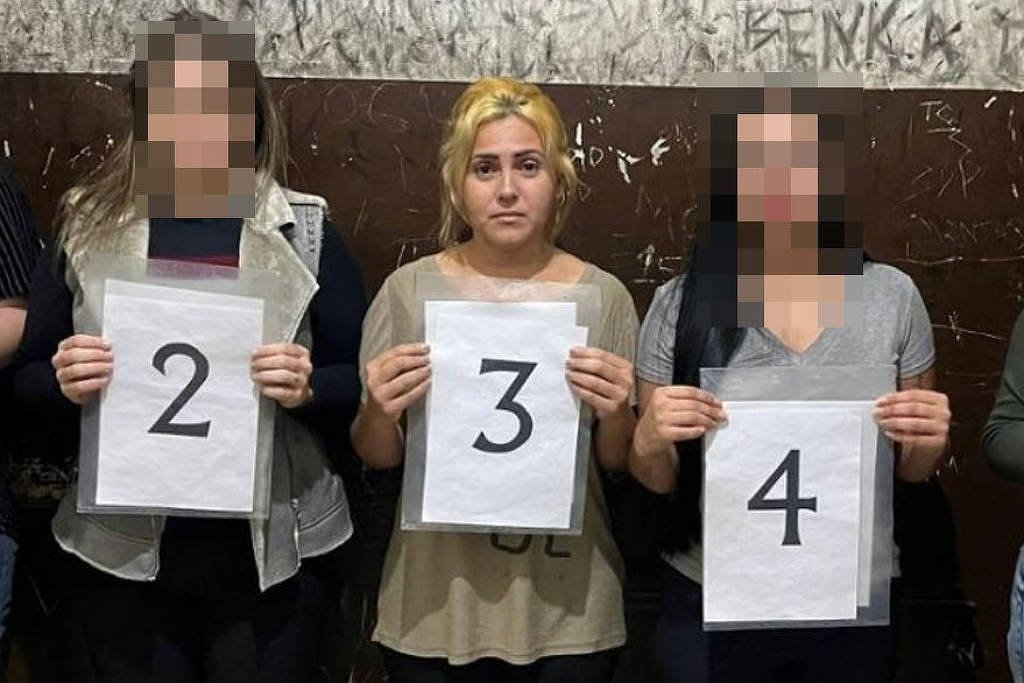 Cinco mulheres em pé, alinhadas lado a lado contra uma parede com marcas e inscrições. Cada uma segura uma folha branca com um número de 1 a 5. A mulher no centro tem cabelo loiro e está descalça, as demais usam roupas casuais e calçados variados.