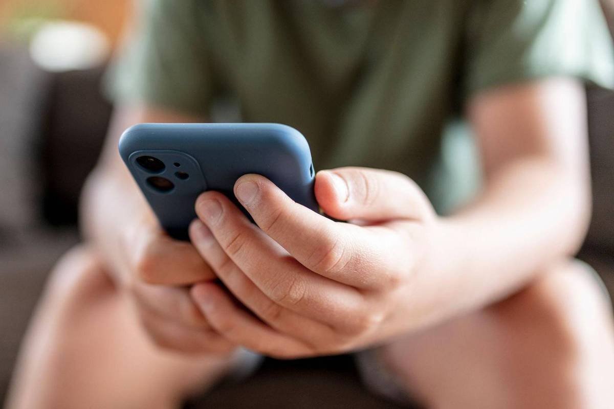 Mãos de uma pessoa seguram um smartphone com capa azul, com foco nas mãos e no aparelho. Fundo desfocado mostra parte do corpo da pessoa e um sofá.