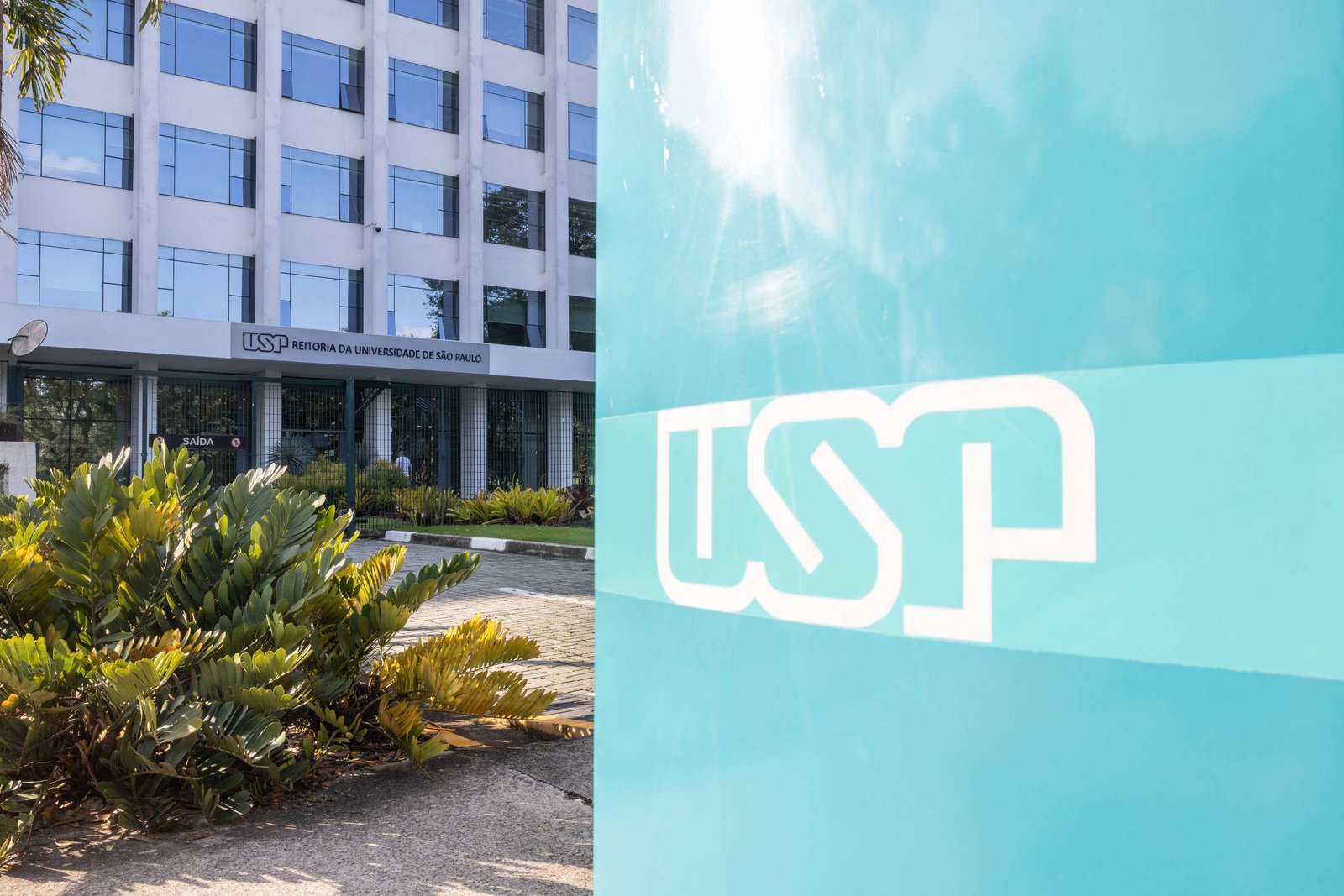 A imagem mostra a fachada de um edifício da Universidade de São Paulo (USP). À esquerda, há um painel azul com o logotipo da USP em destaque. Ao fundo, o edifício possui janelas grandes e uma entrada visível, com vegetação ao redor, incluindo plantas verdes e arbustos.