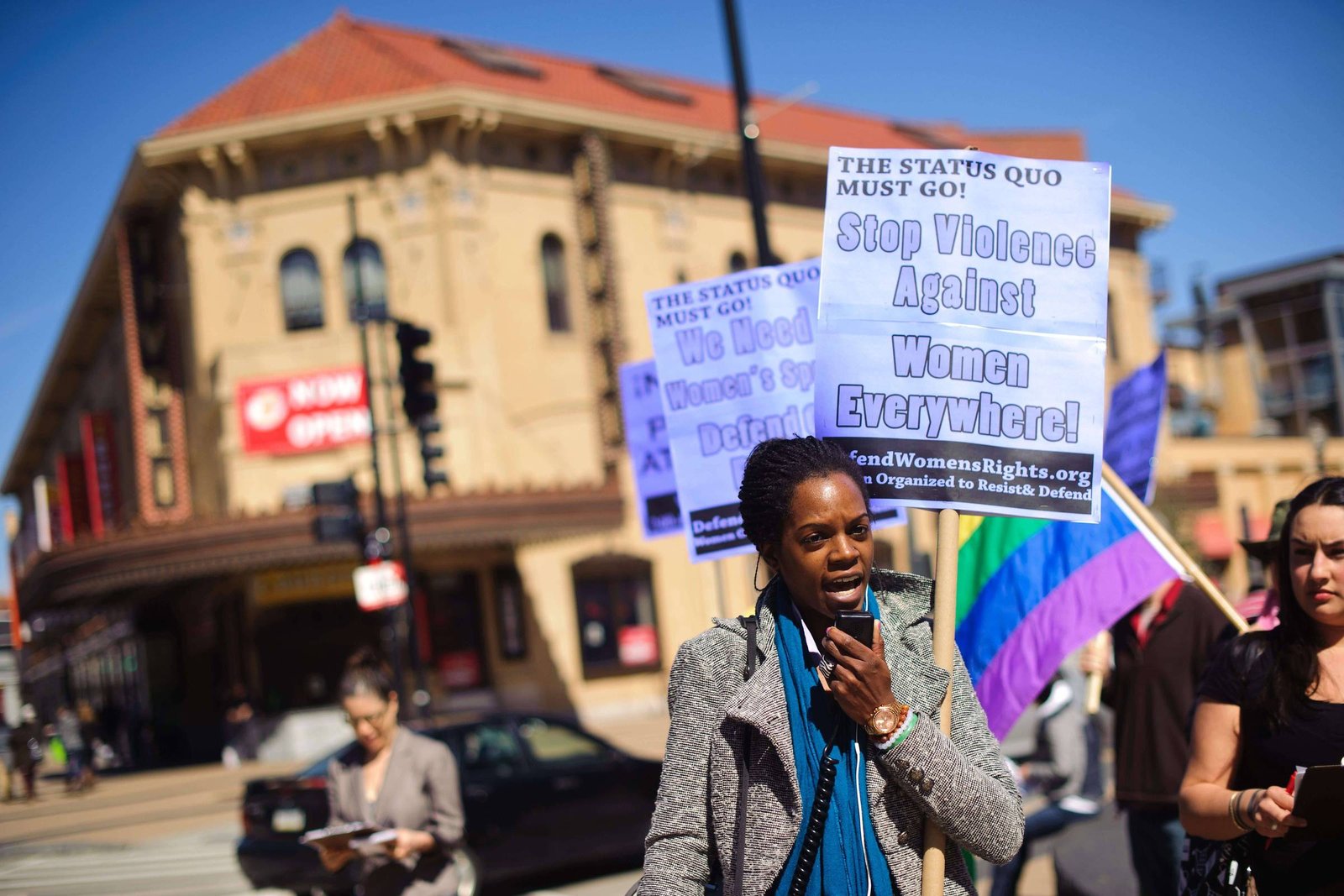 Mulher negra segura microfone e cartaz com texto contra violência a mulheres durante protesto em rua movimentada. Ao fundo, outras pessoas seguram cartazes e bandeira colorida, com prédio antigo e céu azul visíveis.