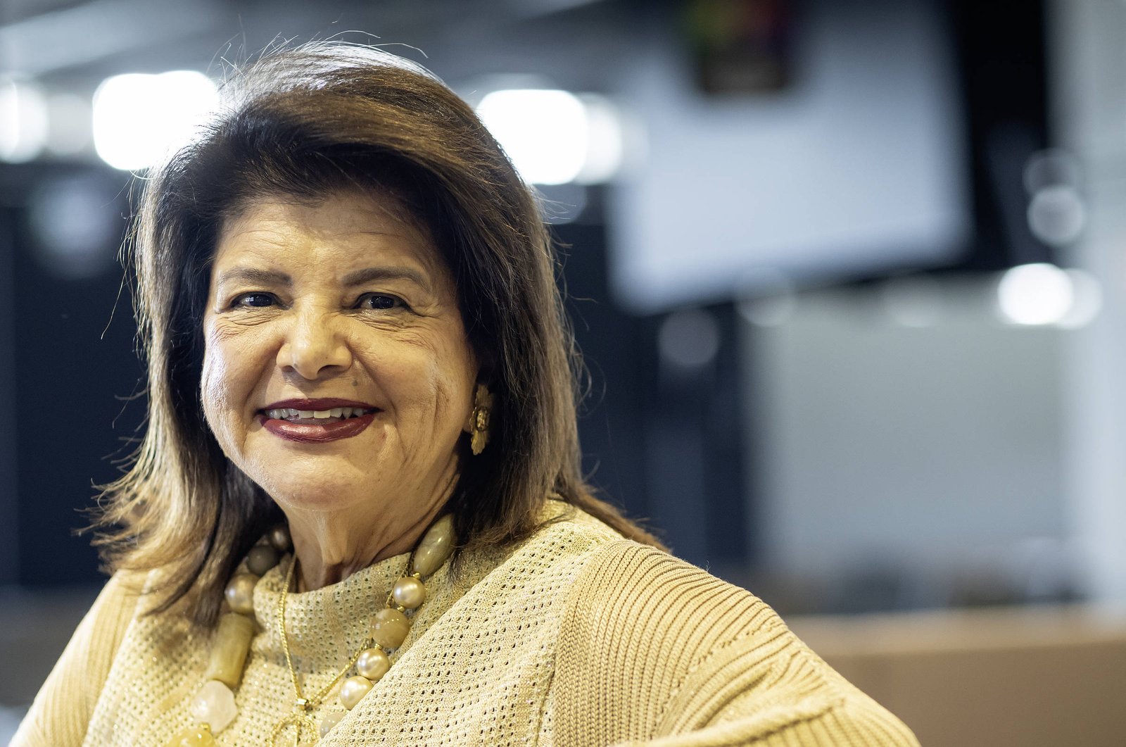 Mulher de meia-idade com cabelo castanho escuro e maquiagem leve sorri para a câmera. Ela veste blusa bege com colar de pérolas e está em ambiente interno com iluminação difusa ao fundo.