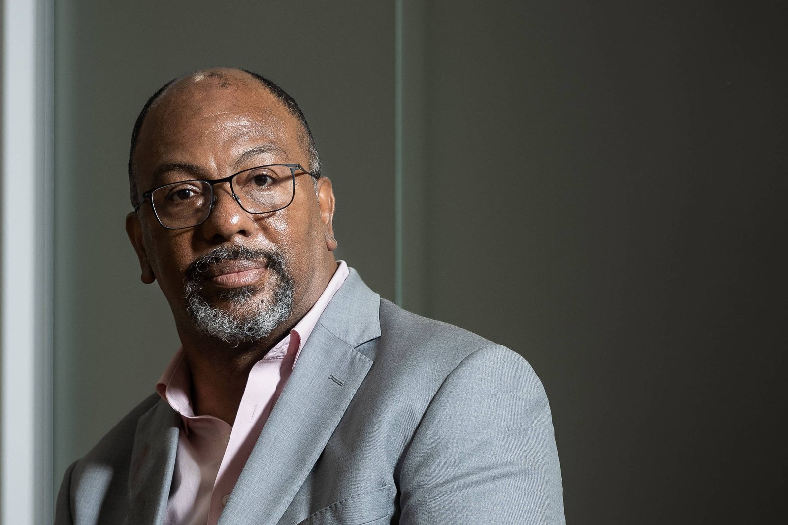 Homem negro de meia-idade sentado em sofá cinza, vestindo terno cinza claro e camisa rosa. Ele usa óculos e olha para frente, com as mãos apoiadas no colo. Fundo neutro e iluminação suave destacam a figura.