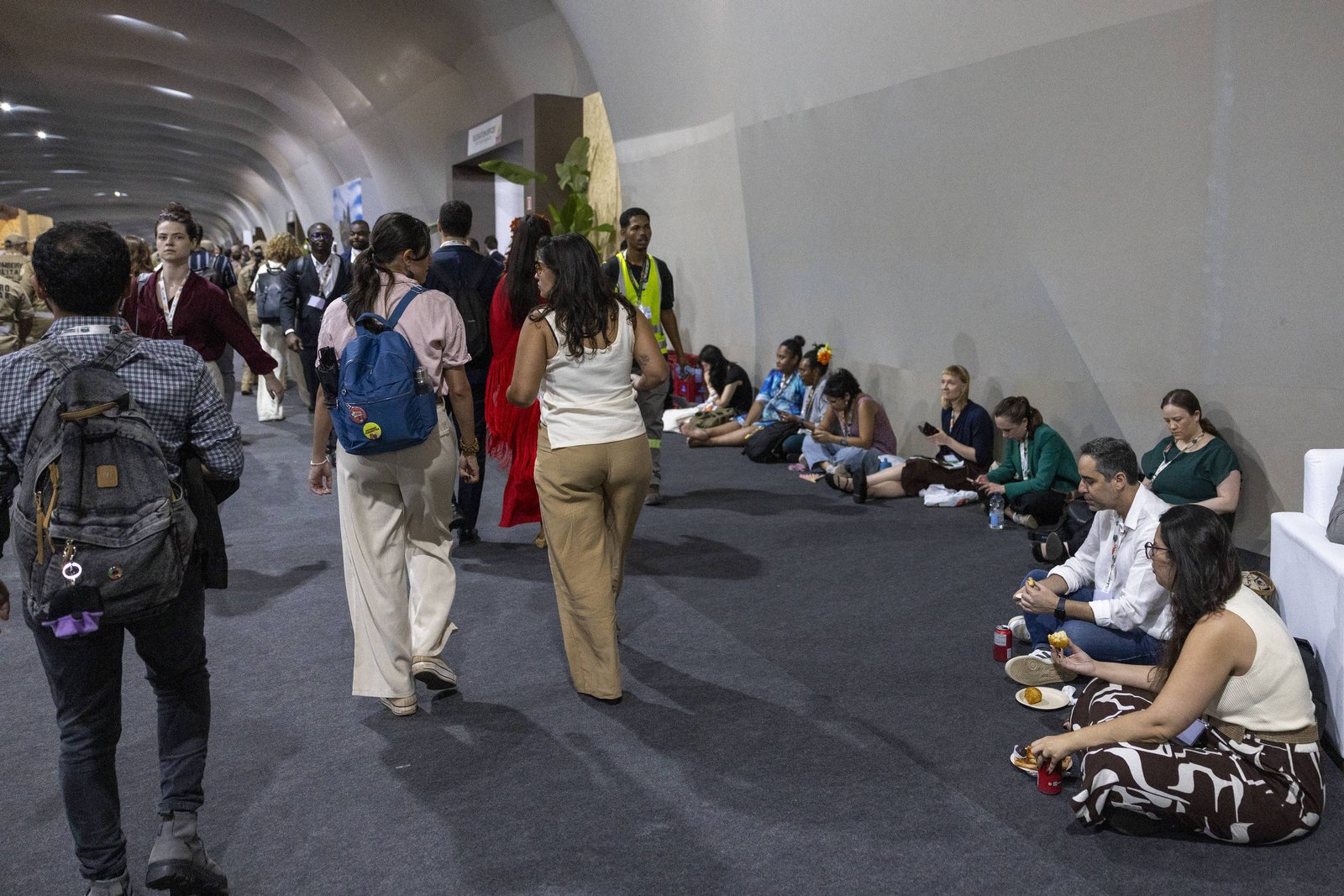 Corredor interno amplo com paredes curvas e iluminação artificial. Pessoas caminham em direção oposta à câmera, algumas com mochilas. À direita, várias pessoas sentadas no chão, algumas comendo, e um homem vestido de terno sentado em sofá branco usando celular.