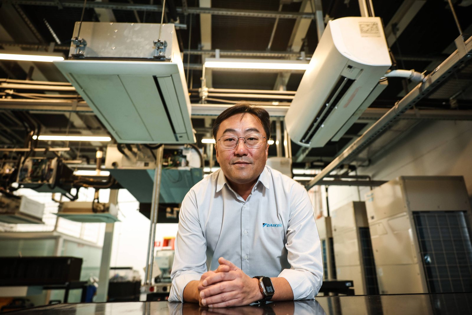 Homem de camisa branca com logotipo encostado em bancada metálica em ambiente industrial. Acima, unidades de ar condicionado e tubulações visíveis no teto.