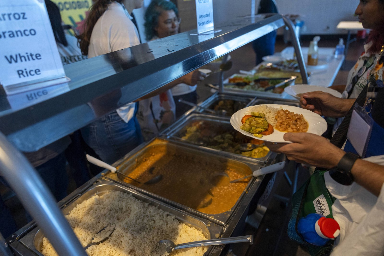 Pessoa segurando prato com comida enquanto se serve em buffet com arroz branco, feijão e outros pratos quentes em bandejas metálicas.