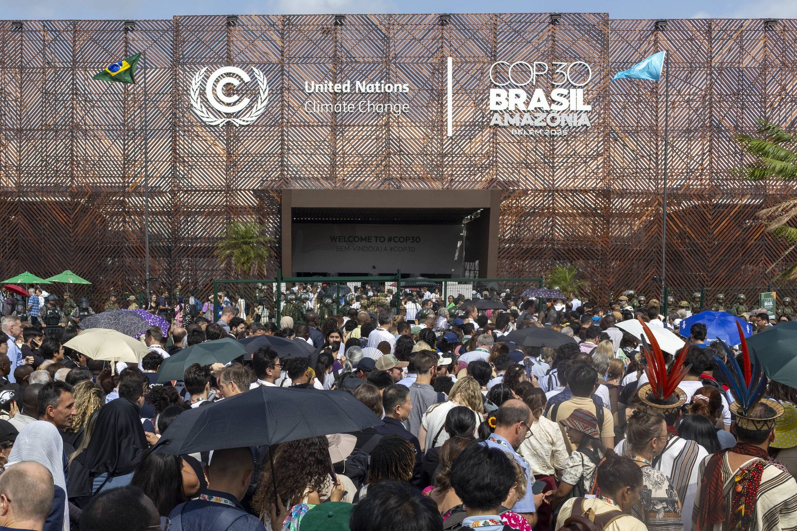 Multidão de pessoas com guarda-chuvas e roupas variadas reunidas em frente à entrada do evento COP30 Brasil Amazônia, com fachada decorada e bandeira do Brasil visível.