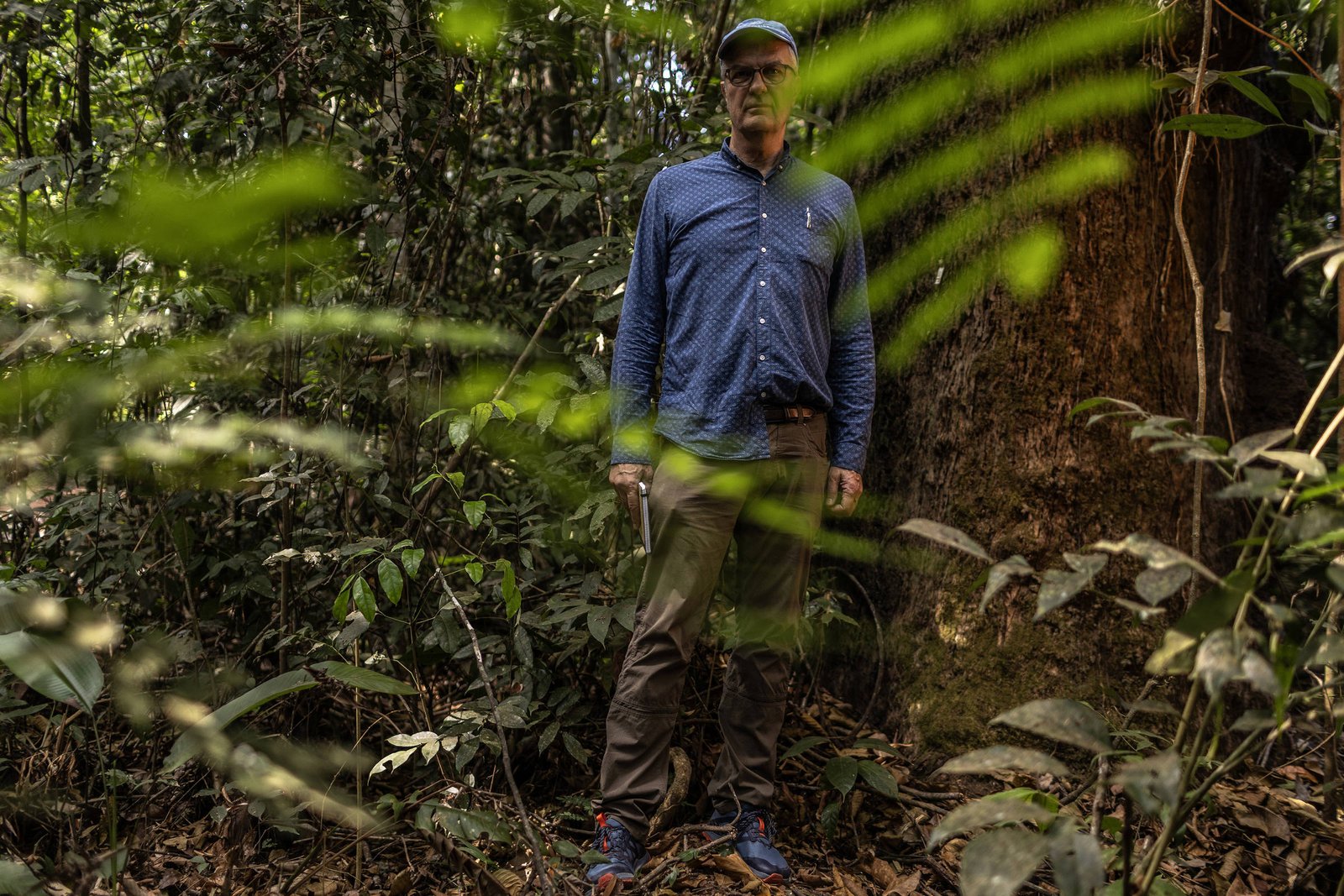 Homem de pé em floresta densa, vestindo camisa azul, calça marrom e boné, segurando uma faca na mão direita. Vegetação verde e tronco de árvore grande ao fundo.
