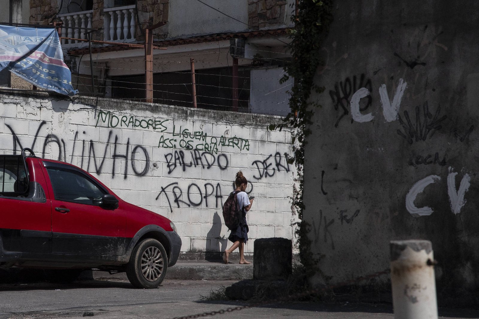Pessoa com mochila caminha ao lado de muro branco coberto por grafites e inscrições em preto. Carro vermelho estacionado à esquerda e edifícios ao fundo. Há várias inscrições com a sigla