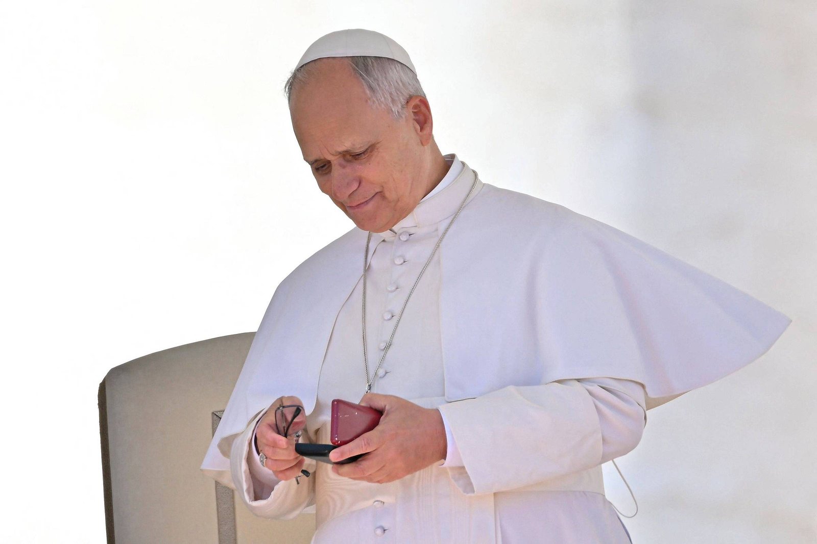 Homem vestido com vestes papais brancas, incluindo solidéu, segura e olha para um dispositivo eletrônico pequeno, possivelmente um celular, com uma capa vermelha. Ele está em pé próximo a uma cadeira cinza, com fundo claro e desfocado.