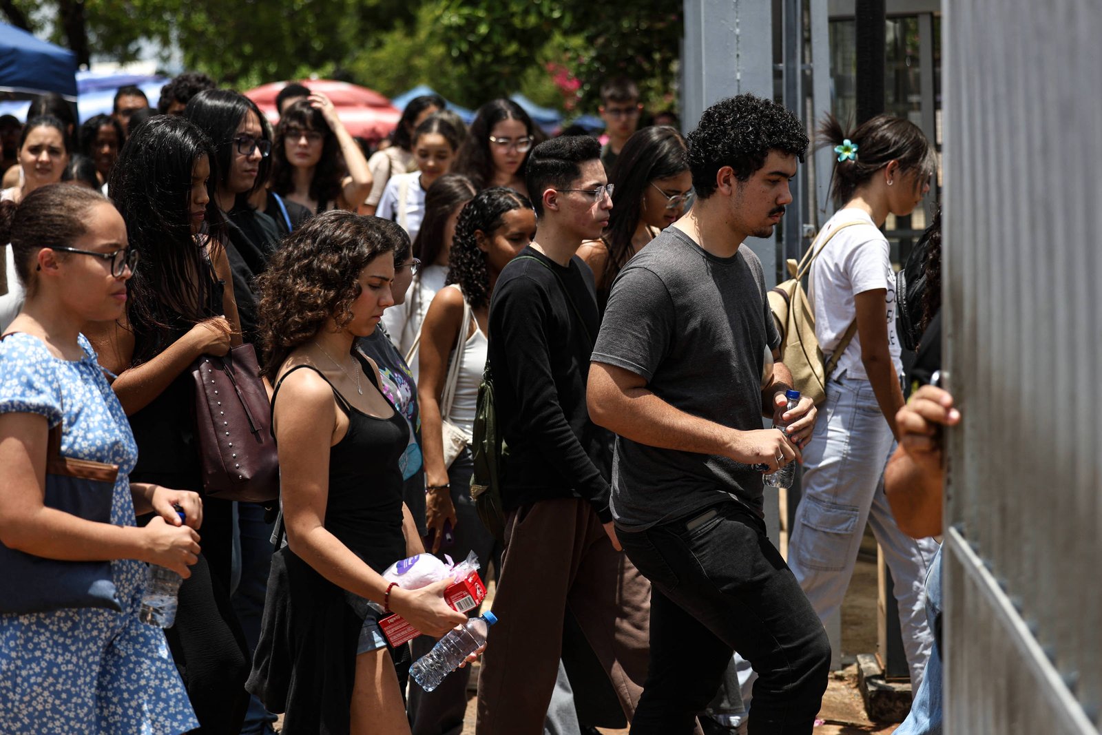 Grupo de jovens em fila ao ar livre, aguardando para entrar por portão metálico. Ambiente com árvores ao fundo e barracas coloridas visíveis.