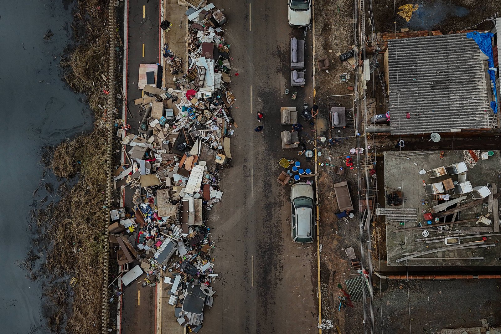 Vista aérea mostra grande quantidade de lixo acumulado ao longo de uma rua estreita entre casas e um rio. Veículos estão estacionados próximos ao lixo, que inclui móveis, entulhos e objetos diversos. Casas com telhados de metal e pessoas circulam pela rua.