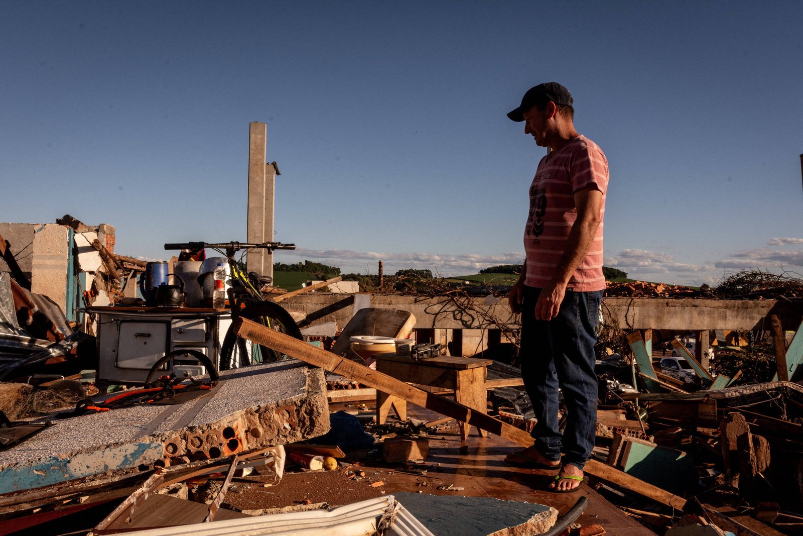Homem de camiseta listrada e boné preto está em pé sobre escombros de uma construção destruída, cercado por pedaços de madeira, metal e móveis danificados sob céu azul claro.
