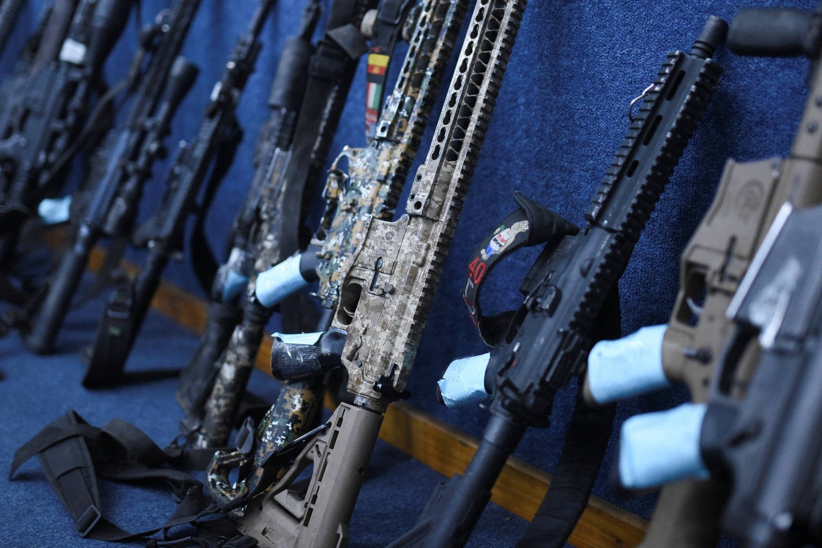 Diversos rifles automáticos alinhados encostados em uma parede azul, com partes das armas cobertas por fitas azuis.