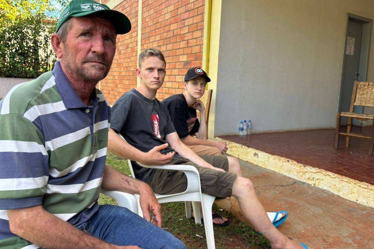 Três homens sentados em cadeiras plásticas na área externa de uma casa com parede de tijolos à vista. O homem mais velho, à esquerda, usa boné verde e camisa listrada, olhando para a câmera. Os dois homens mais jovens, ao centro e à direita, vestem camisetas escuras e bermudas, um deles usa chinelos azuis. Ao fundo, uma porta aberta e uma cadeira de madeira estão visíveis.