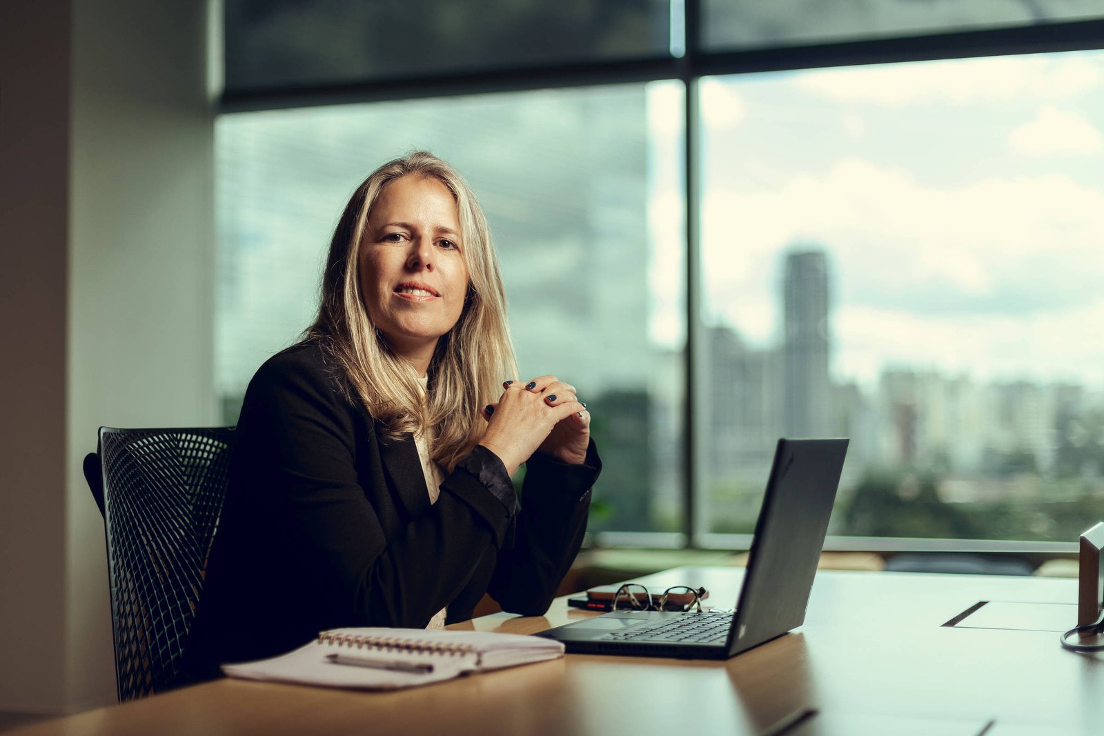 Mulher loira de meia-idade sentada à mesa de escritório com laptop aberto, caderno e óculos. Janela grande ao fundo mostra prédios e céu nublado.