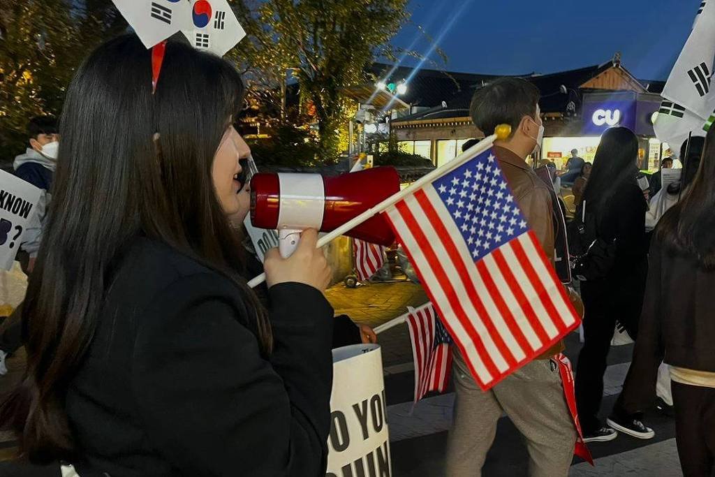 Grupo de pessoas em manifestação noturna segurando bandeiras dos Estados Unidos e Coreia do Sul. Mulher com megafone e rosto pintado participa do protesto em área urbana com iluminação artificial.