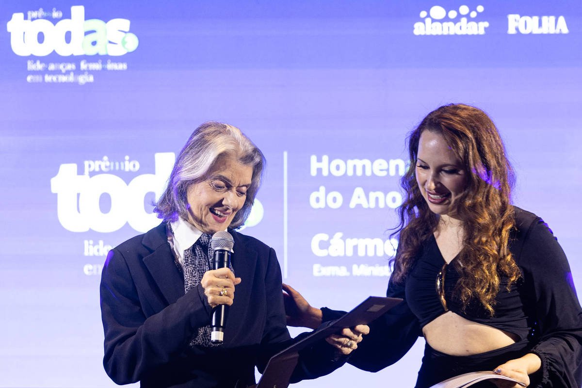 Duas mulheres no palco durante cerimônia. A mulher à esquerda segura microfone e documento, a da direita sorri e segura outro documento. Fundo azul com texto de premiação visível.