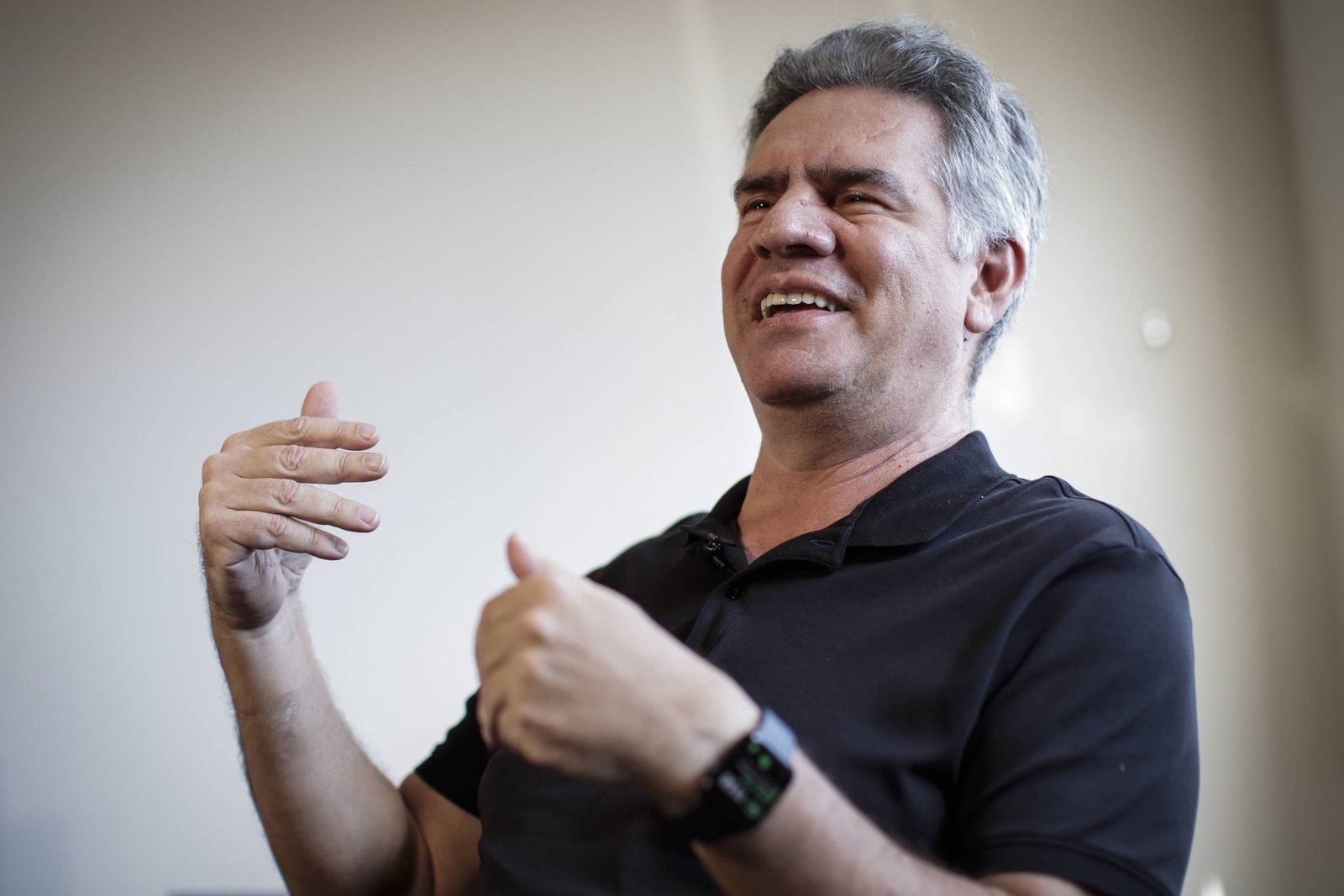Homem de meia-idade com cabelo grisalho veste camisa preta e usa relógio inteligente no pulso esquerdo. Ele está sentado em ambiente interno com parede clara ao fundo, gesticulando com as mãos enquanto fala.