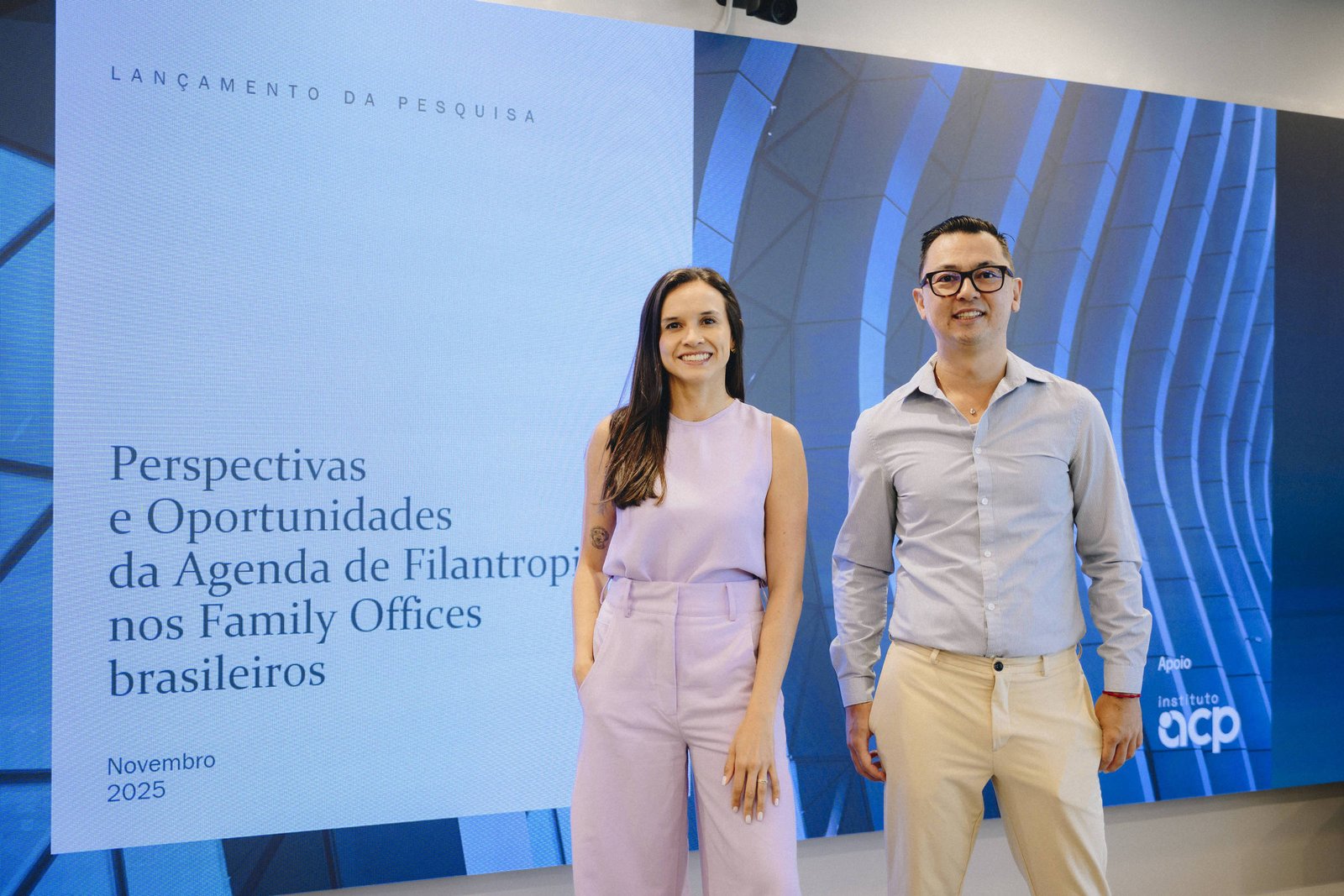 Dois profissionais posam em pé diante de tela grande que exibe o título da pesquisa