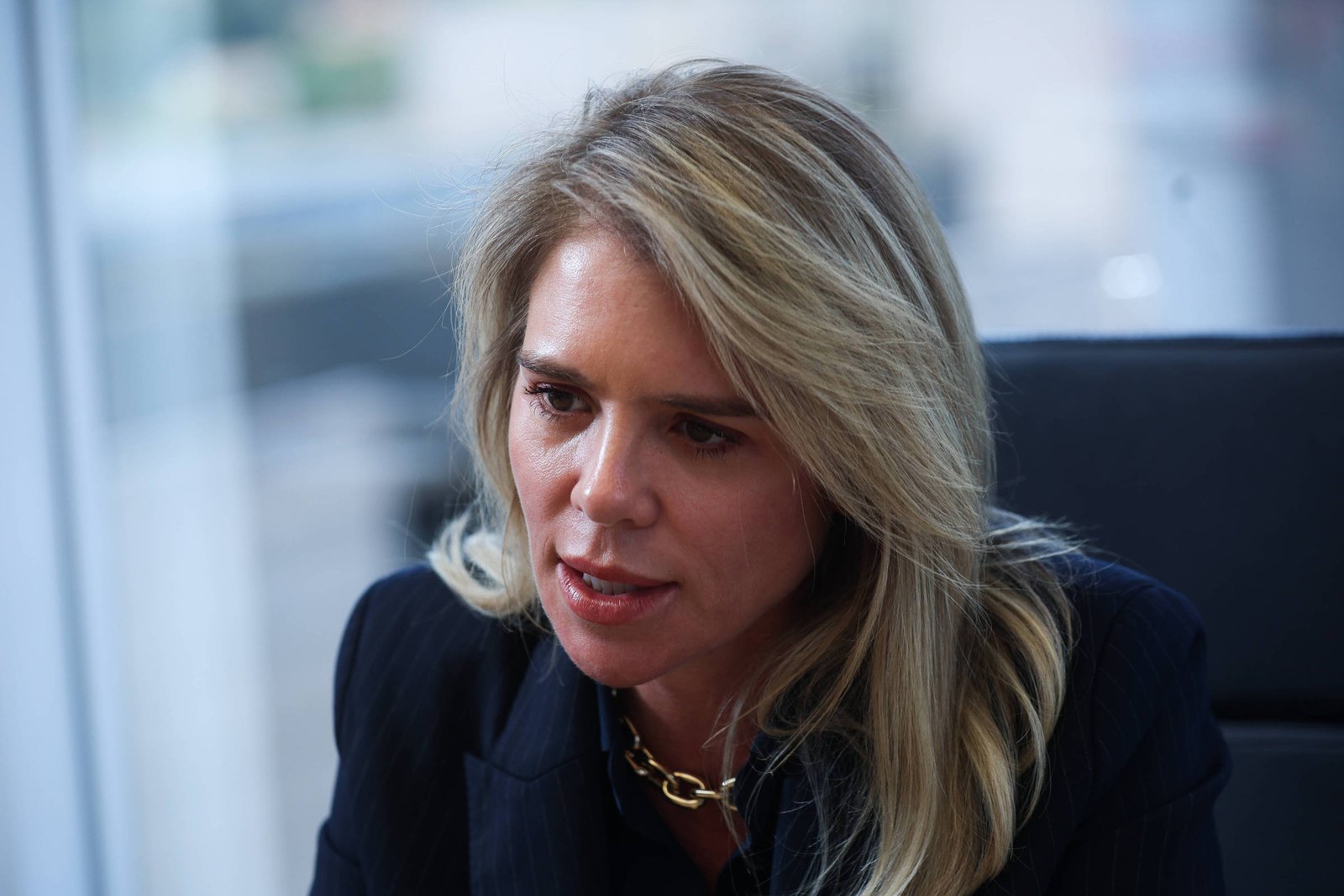 Mulher loira com cabelo solto, vestindo blazer preto e colar dourado, sentada em cadeira preta em escritório com janela ao fundo.