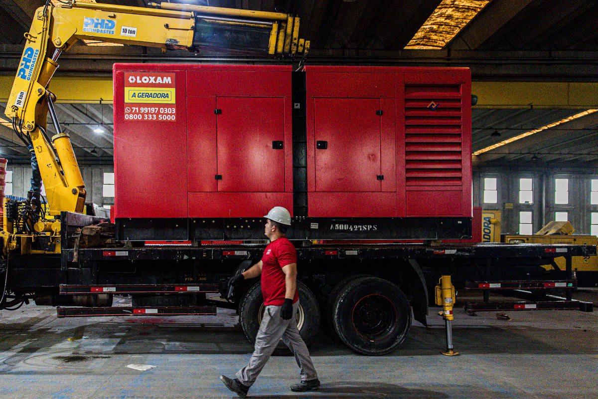 Homem com capacete branco e camiseta vermelha caminha em galpão industrial à frente de gerador vermelho montado sobre caminhão. Guindaste amarelo está posicionado ao lado do gerador, com iluminação artificial no teto do galpão.