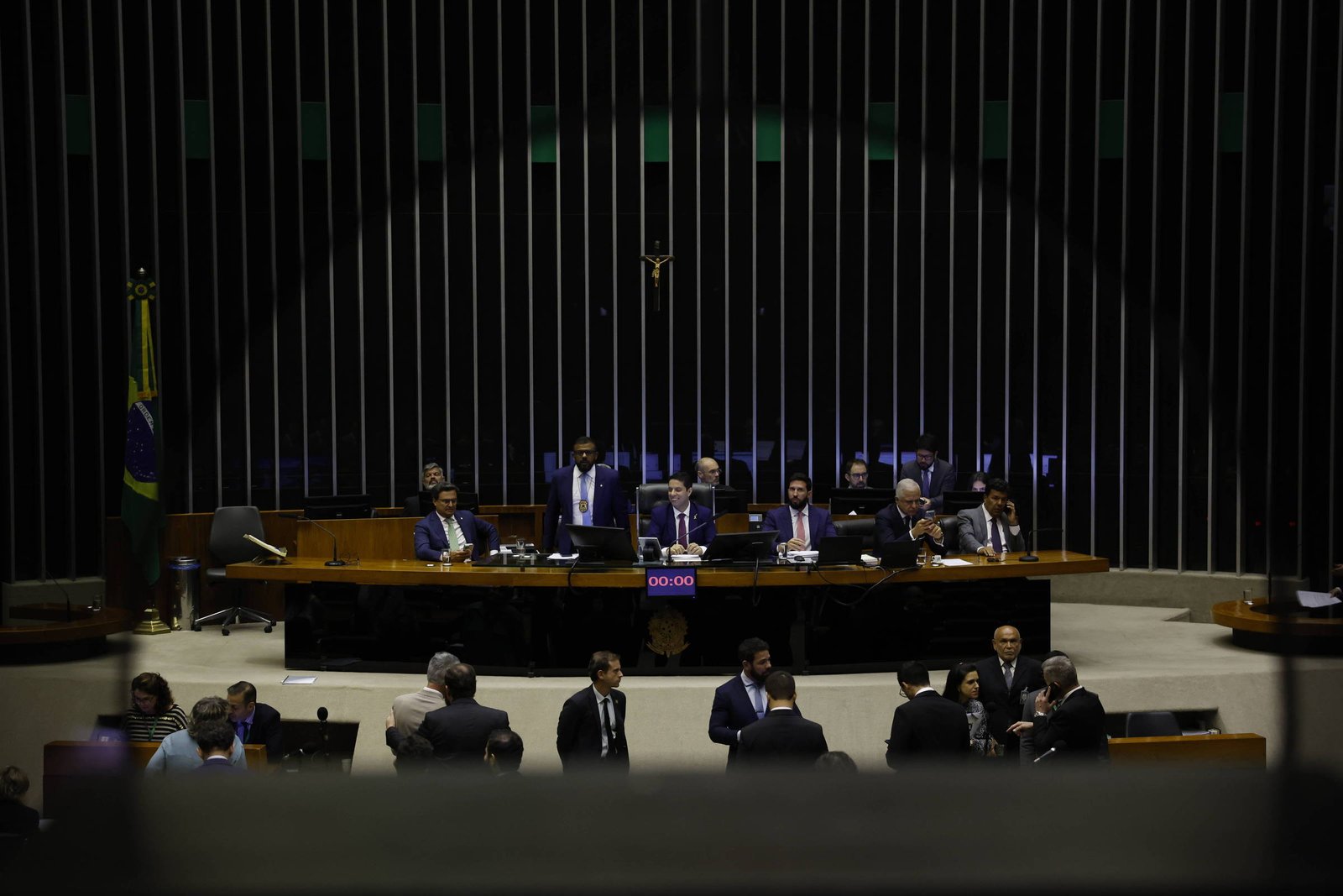 Deputados sentados e em pé no plenário da Câmara dos Deputados, com mesa principal ao fundo e bandeira do Brasil à esquerda. Ambiente formal com iluminação baixa e estrutura arquitetônica com barras verticais.