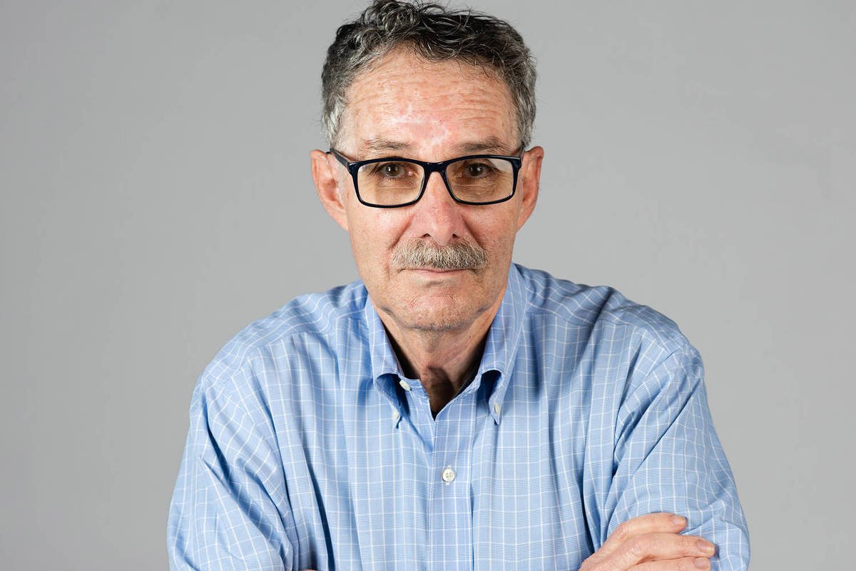 Homem de meia-idade com cabelo grisalho e óculos escuros, veste camisa azul clara de manga longa e está com os braços cruzados, posando contra fundo cinza neutro.