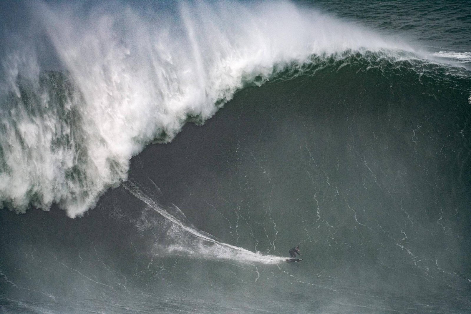 Surfista desliza em uma onda gigante no mar, com a crista da onda formando espuma branca acima dele.