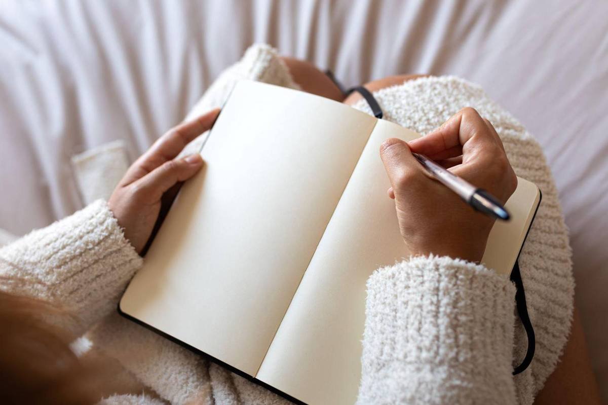 Pessoa com suéter de tricô claro segura caneta e escreve em caderno aberto com páginas em branco, sentada em superfície macia e clara.
