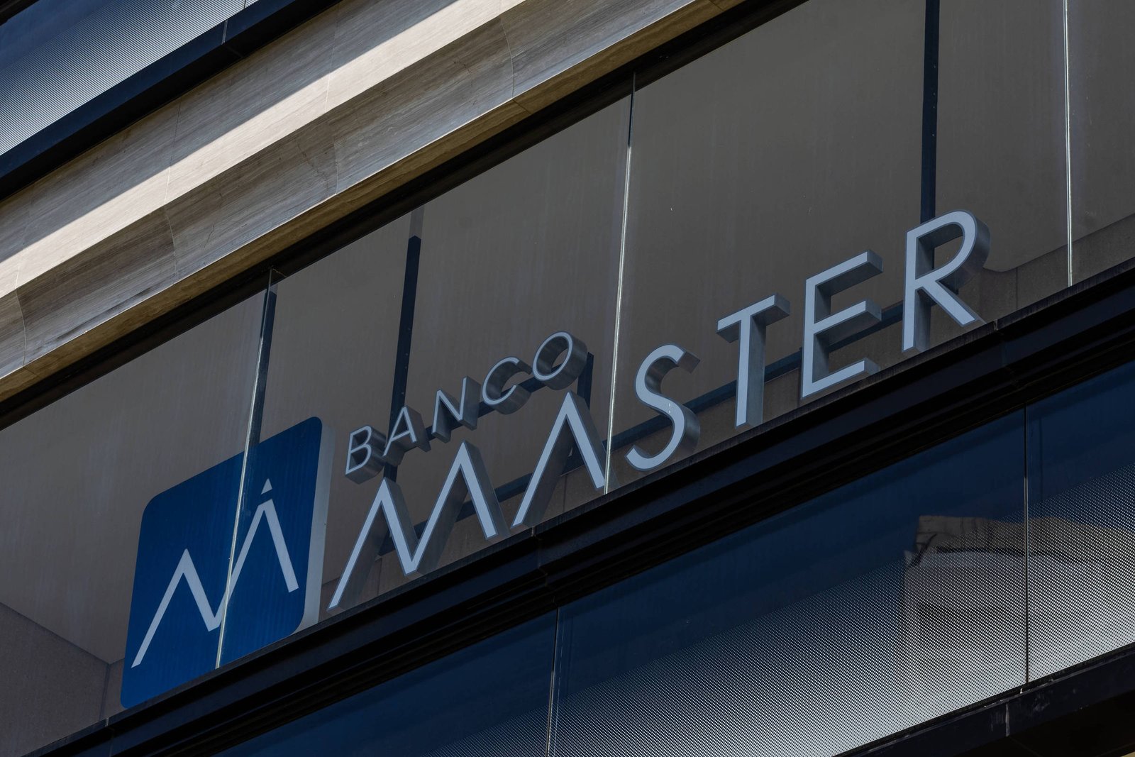 Fachada de vidro de agência do Banco Master com logo azul e branco e nome em letras metálicas fixadas na parede externa.