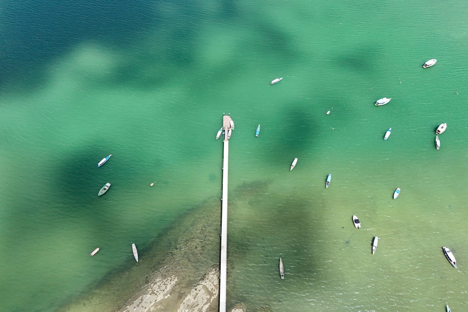 A imagem mostra uma vista aérea de um pier que se estende sobre águas de tonalidade verde e azul. Vários barcos estão ancorados ao redor do pier, e a água é clara, permitindo ver o fundo. A costa é visível à esquerda, com uma área de areia e rochas.
