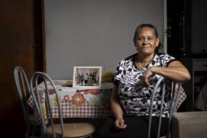 Mulher de meia-idade sentada em cadeira de metal ao lado de mesa com toalha xadrez e floral. Na mesa, quadro com foto de grupo familiar. Ambiente interno com parede cinza e porta de madeira ao fundo.