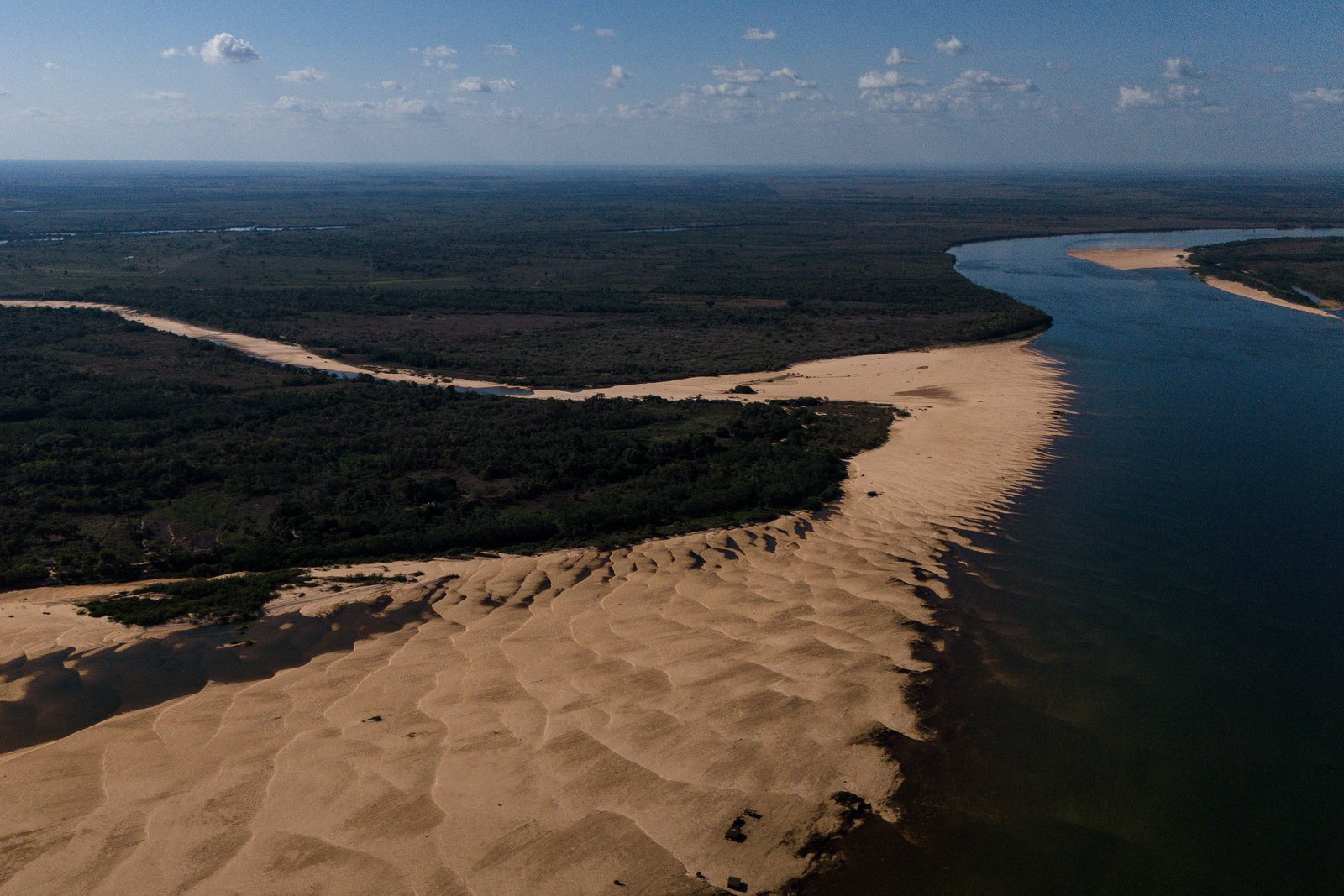 Rio largo contorna faixa extensa de dunas de areia clara que se estende até área de vegetação densa e escura. Céu azul com poucas nuvens ao fundo.