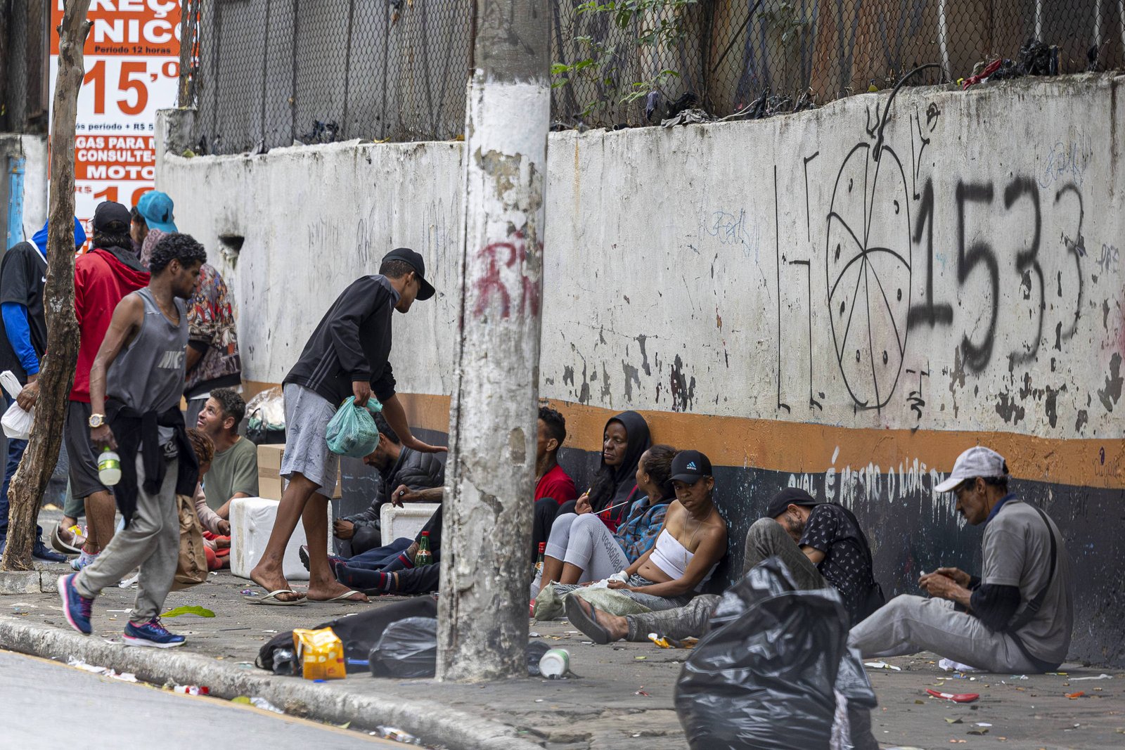 Grupo de pessoas sentadas e em pé na calçada ao lado de muro pichado com grafite e números. Alguns carregam sacolas plásticas e objetos pessoais. Ambiente urbano com lixo e entulho no chão.