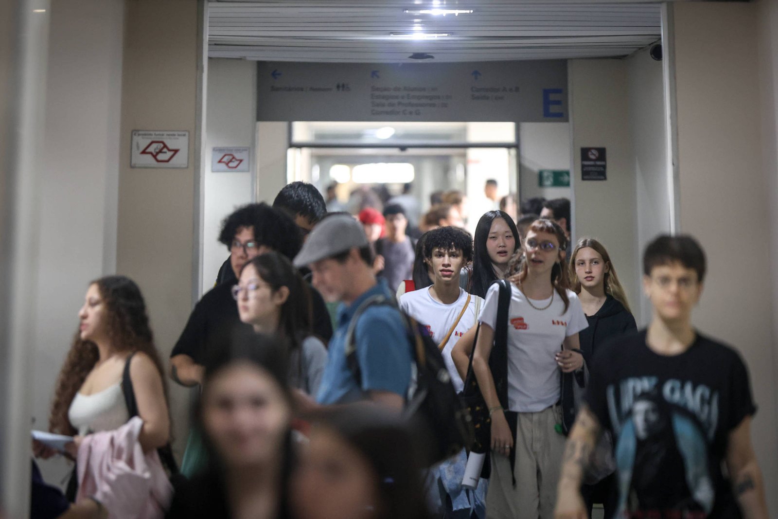 Grupo diversificado de pessoas caminha por corredor interno com iluminação artificial. Algumas carregam mochilas e bolsas, e há placas indicativas nas paredes.