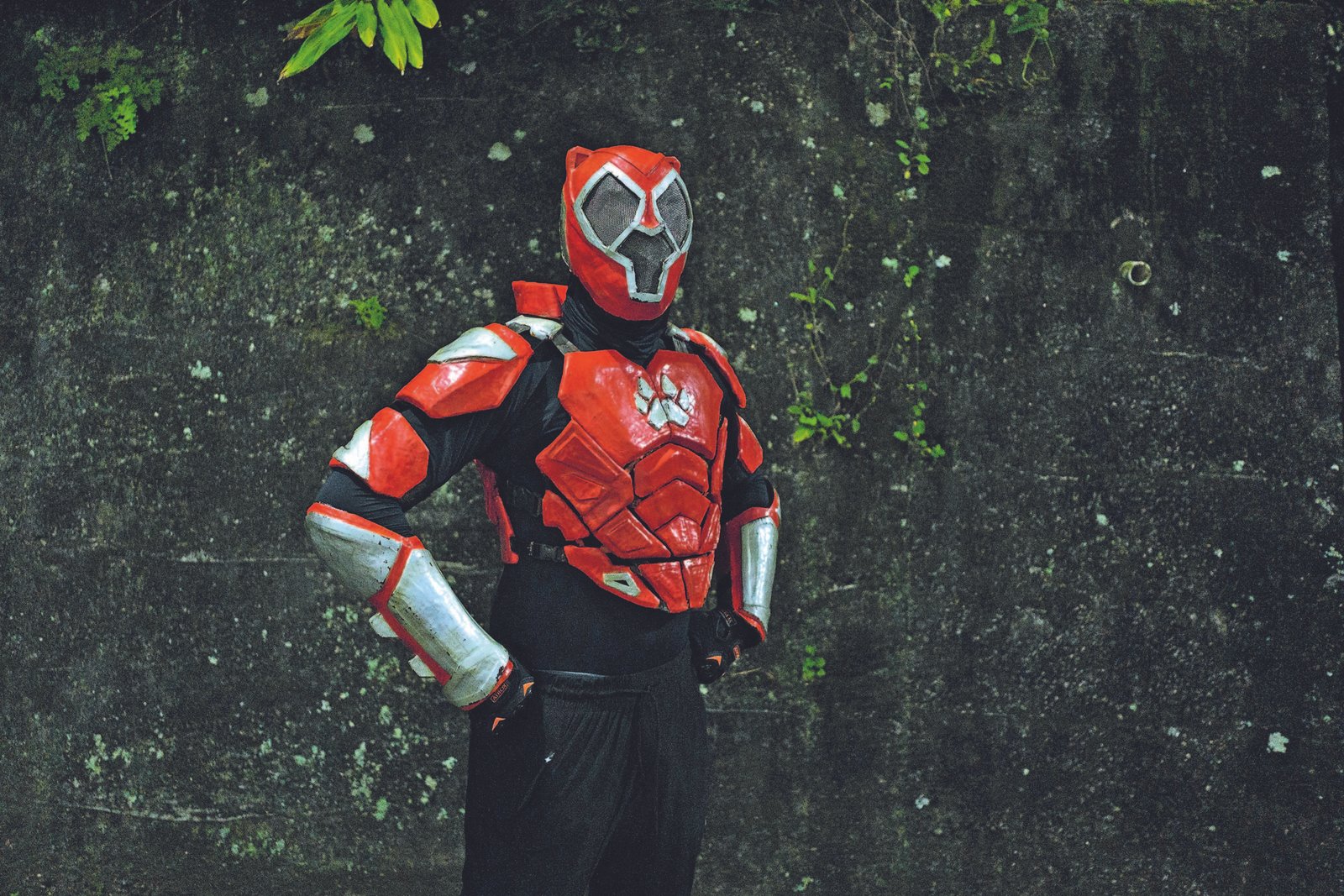 Indivíduo veste fantasia vermelha e preta com armadura e máscara, posicionado com as mãos na cintura diante de parede escura com vegetação.