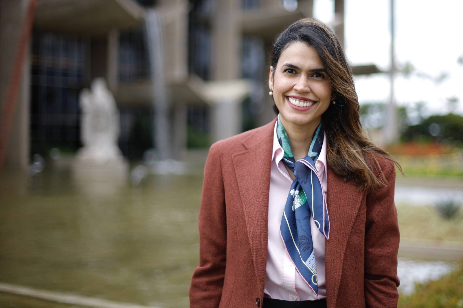 Mulher jovem com cabelo castanho solto, vestindo blazer marrom, camisa clara e lenço azul no pescoço, sorri para a câmera. Ao fundo, fonte com estátua desfocada e vegetação em área urbana.