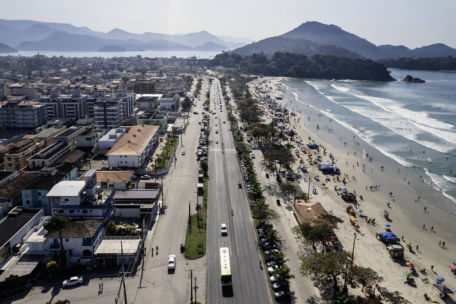 Avenida com tráfego moderado separa área urbana de praia cheia de pessoas e barracas. Montanhas e mar ao fundo sob céu claro.