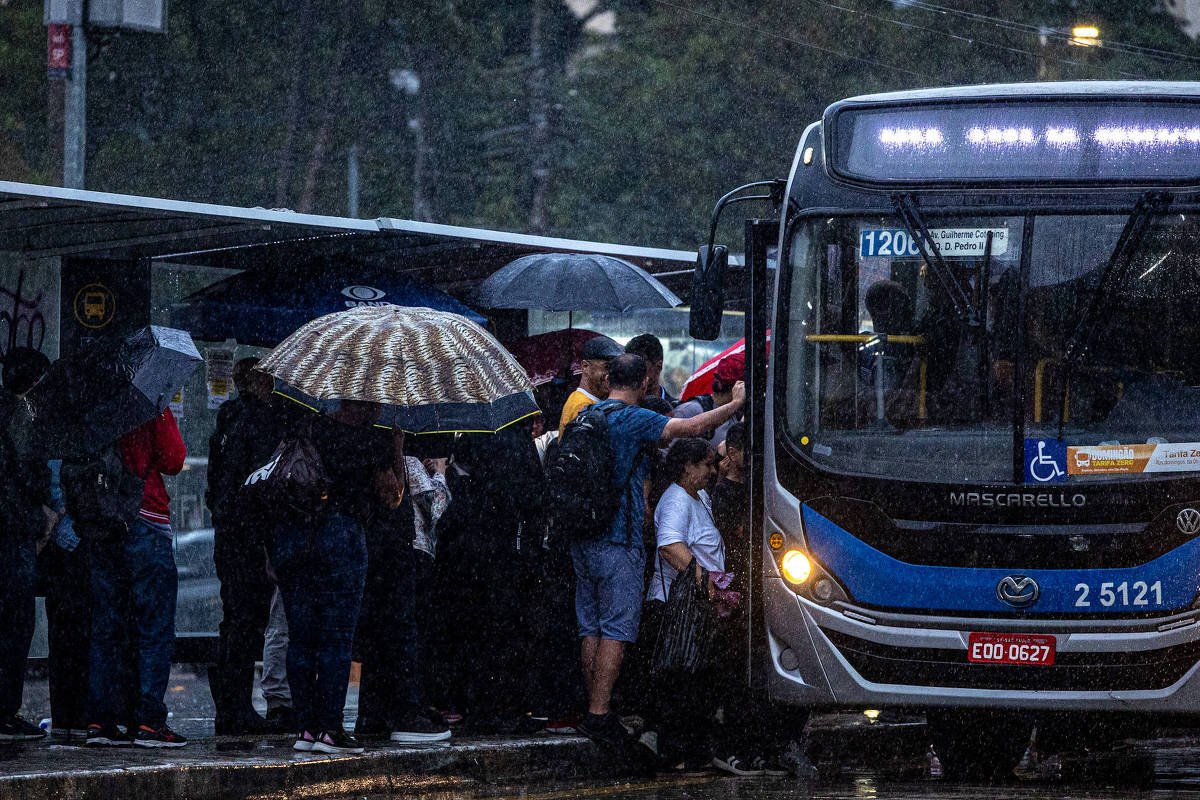 Grupo de pessoas aguarda e embarca em ônibus na parada durante chuva intensa. Vários usam guarda-chuvas e roupas escuras, e o ônibus exibe placa de acessibilidade.