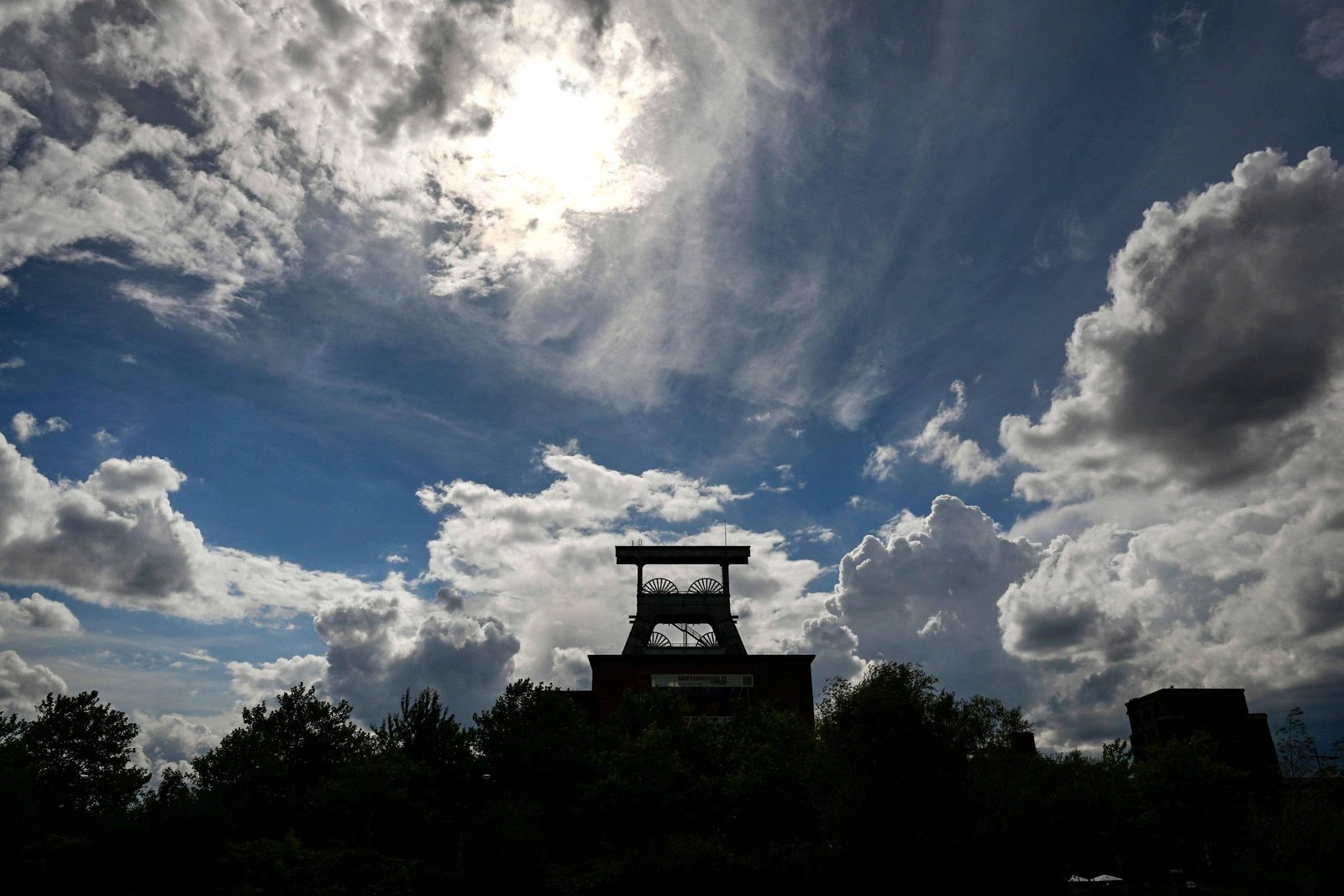 Silhueta de estrutura industrial com torre metálica e edifício ao fundo, cercada por árvores. Céu azul com nuvens densas e sol parcialmente encoberto.