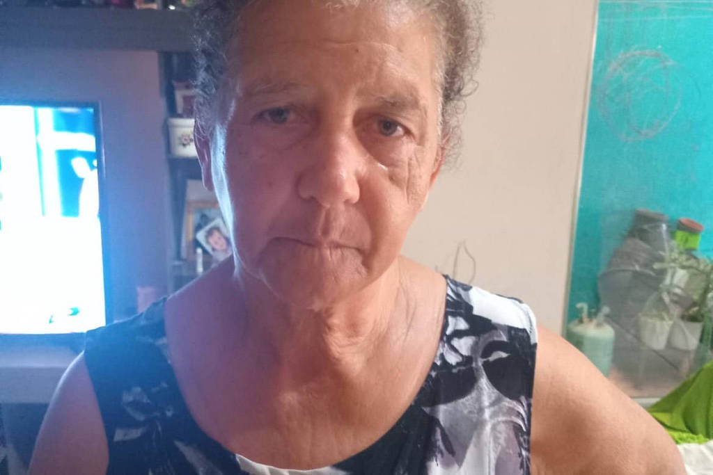 Mulher de meia-idade com cabelo preso, vestindo blusa preta e branca, está em ambiente interno com parede azul ao fundo e objetos ao redor.