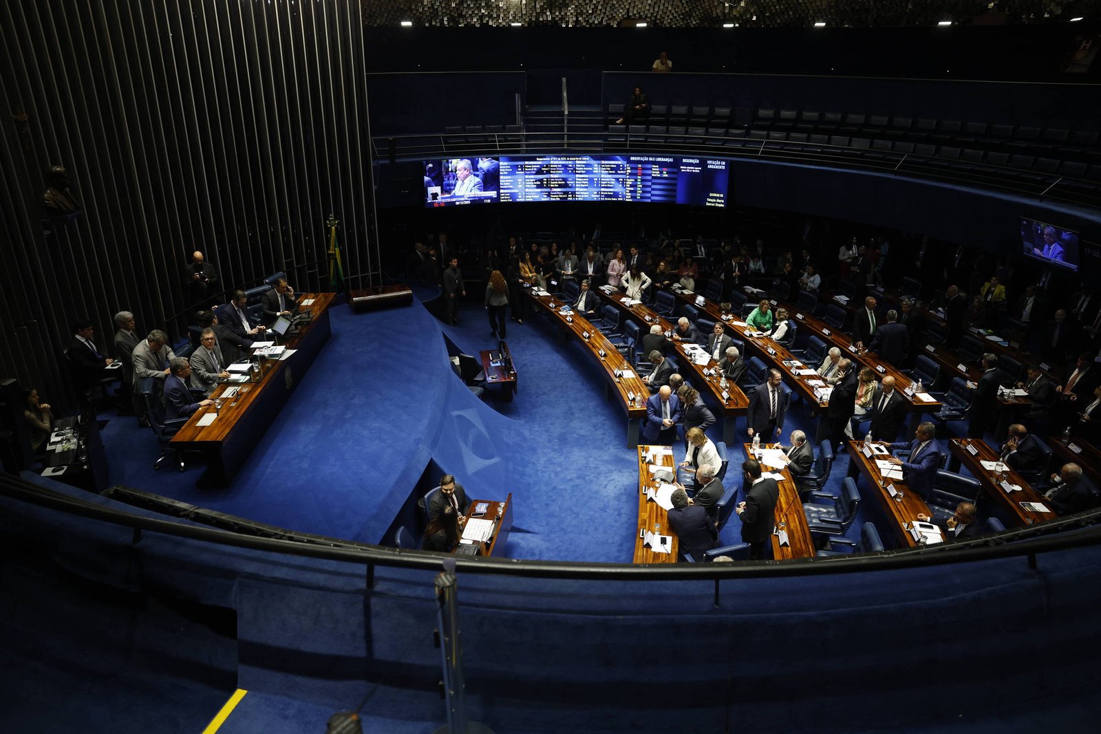 Senadores sentados em seus lugares no plenário do Senado Federal, com mesas de madeira e cadeiras azuis. Painéis eletrônicos exibem informações ao fundo, e o ambiente é iluminado artificialmente.