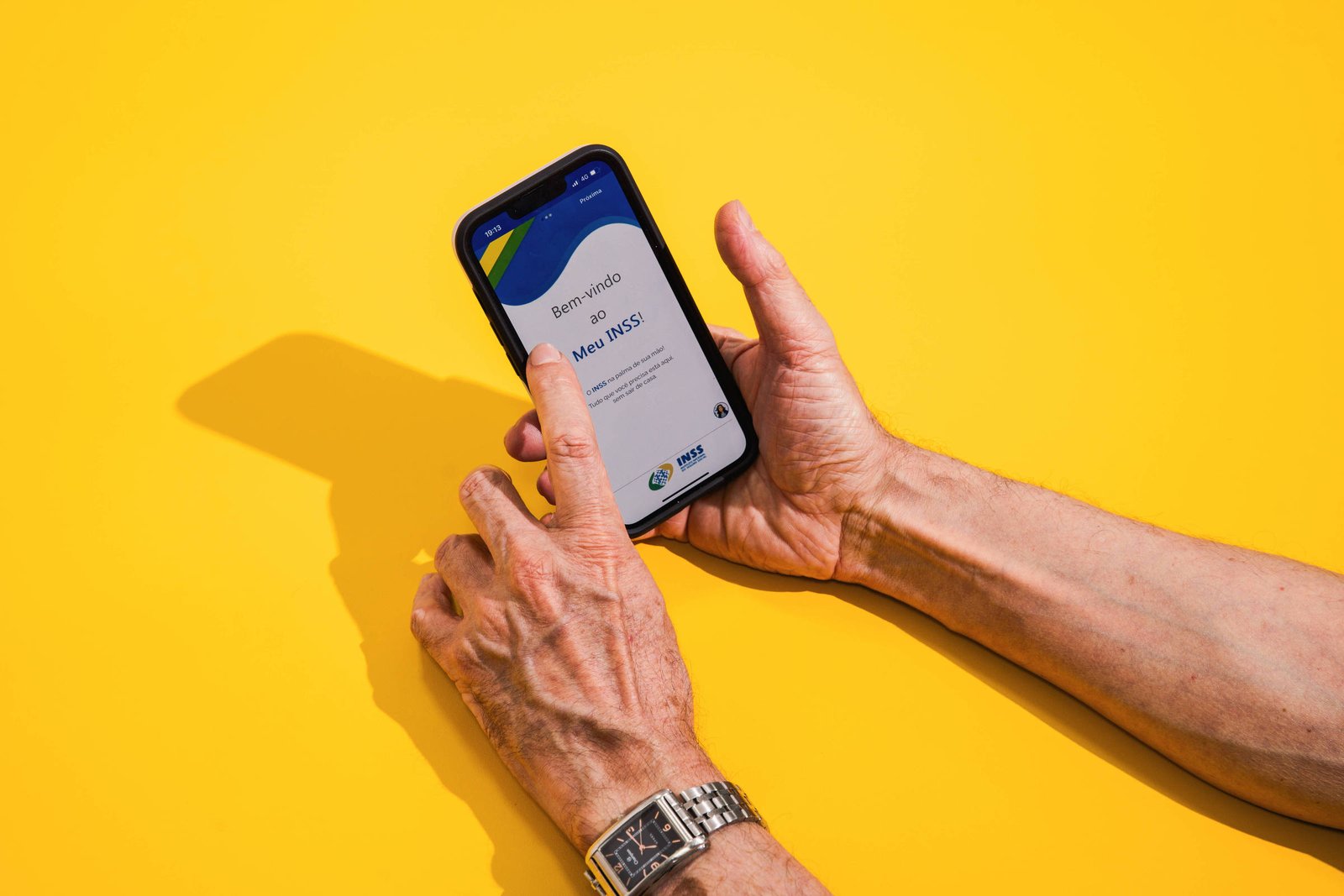 A imagem mostra uma mão segurando um smartphone com a tela voltada para cima. O smartphone exibe uma interface de aplicativo, possivelmente relacionada a serviços financeiros, com um fundo azul e verde. A mão é de uma pessoa idosa, e um relógio de pulso prateado é visível no pulso. O fundo da imagem é amarelo.